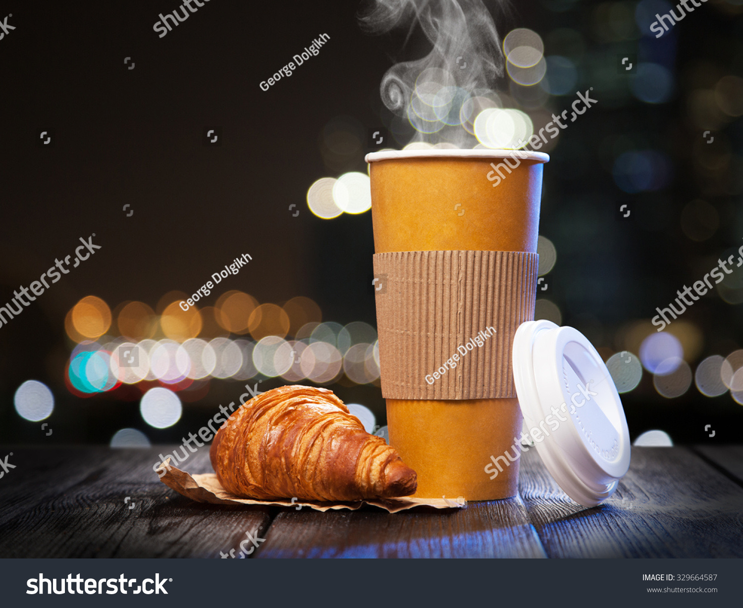 Takeaway coffee in a paper cup with a croissant against a bokeh city night background